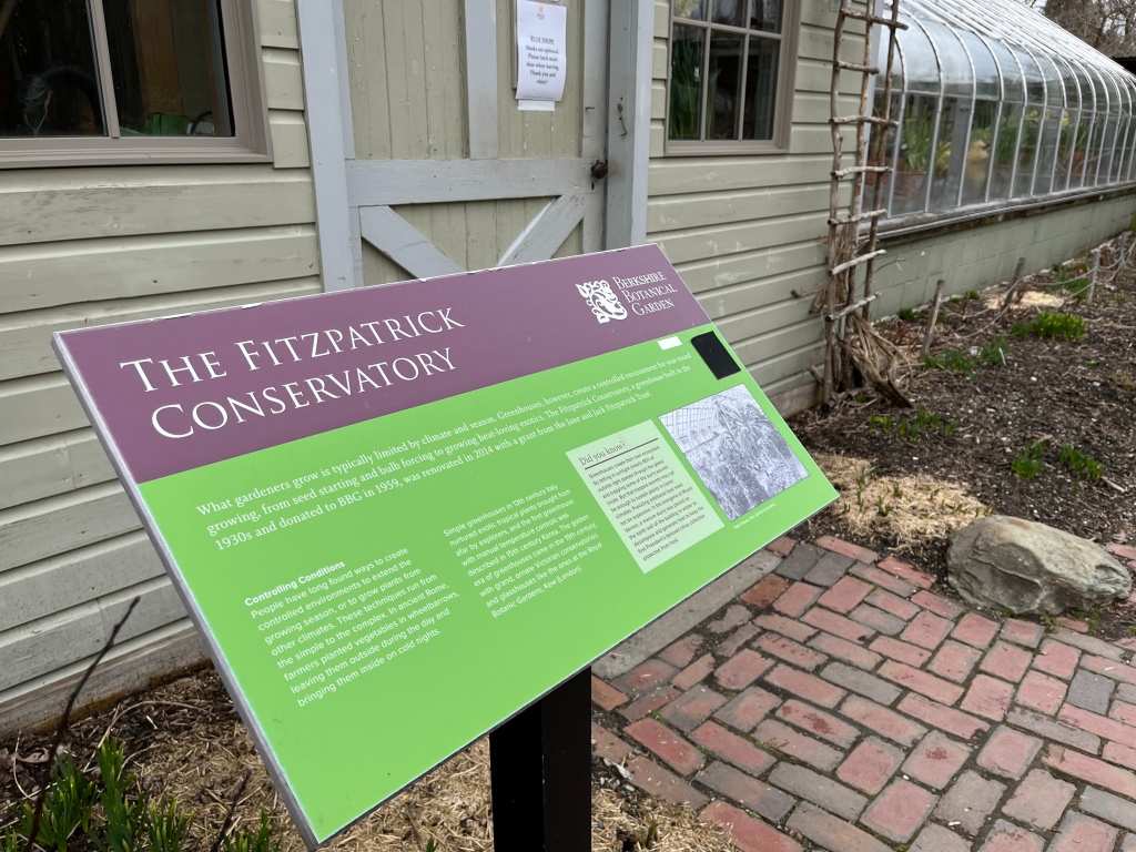 A green and purple sign in front of a greenhouse that reads the Fitzpatrick Conservatory